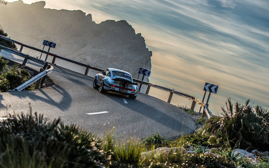 Vintage car navigating a winding mountain road at sunset with dramatic cliffs in the background.